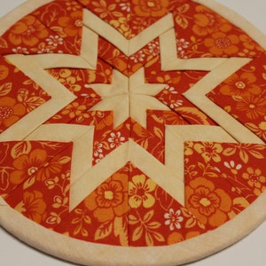 May include: A circular fabric wall hanging featuring a folded star pattern. The design combines cream-colored fabric strips with a vibrant orange and red floral print fabric. The entire piece is framed within a circular wooden embroidery hoop.