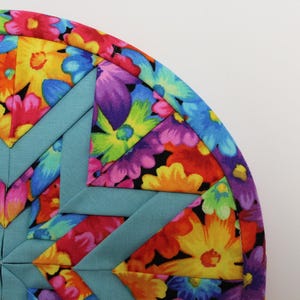 May include: A circular quilted potholder featuring a vibrant floral pattern with red, yellow, orange, purple, and blue flowers. The design incorporates a geometric star motif constructed from solid teal fabric strips, finished with a matching teal border.