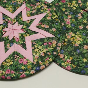 May include: Two round quilted pot holders. One features a pink star design on a dark green floral background. The other pot holder has the same floral pattern. The floral pattern includes pink, yellow, and blue flowers.