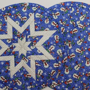 May include: Two blue, round placemats with a white star design in the center. The background is covered in a pattern of snowmen and snowflakes. The star is made of white fabric with a small gray dot pattern.