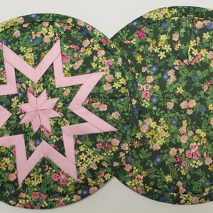 May include: Two round quilted pot holders. One features a pink star design on a dark green floral background. The other pot holder has the same floral pattern. The floral pattern includes pink, yellow, and blue flowers.