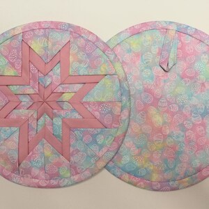 May include: Two round quilted pot holders. One features a pink star design on a pastel egg-patterned background. The other has the same egg pattern with a loop for hanging. The pot holders are approximately 8 inches in diameter.
