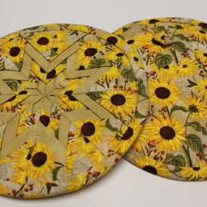 May include: Two round, quilted pot holders with a sunflower pattern. One pot holder features a star design in the center. The other pot holder has a full sunflower print with green leaves and red berries. The background is a light beige.