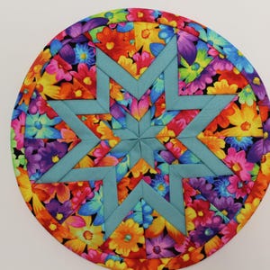 May include: A circular quilted potholder featuring a central eight-pointed star in solid teal fabric. The surrounding background consists of a vibrant floral pattern with red, yellow, purple, and blue flowers on a black fabric base.