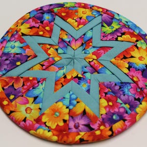 May include: A circular quilted potholder featuring a vibrant floral fabric with red, yellow, purple, and blue flowers. The center displays a prominent eight-pointed star design constructed from solid light blue fabric segments.