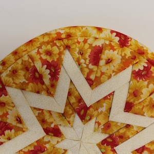 May include: A round, quilted hot pad with a starburst design. The fabric is yellow with red and white floral print. The center of the starburst is white.
