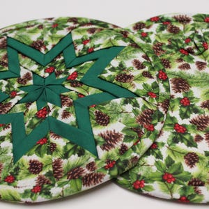 May include: Two round Christmas-themed quilted placemats. One features a green star design, while both have a festive pattern of pinecones, holly, and red berries on a white background. These placemats are perfect for holiday decor.