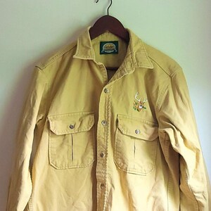 May include: A yellow long-sleeved button-down shirt with a deer embroidered on the front. The shirt has a green label with the text "The Great Outdoors" on it.