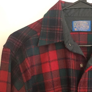 May include: A red, black, and green plaid button-down shirt with a black collar. The shirt has a chest pocket and a label that reads "Pendleton". The shirt is hanging on a black hanger.