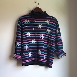 Könnte beinhalten: Schwarzer gestreifter Pullover mit Stehkragen und Schneeflocken-Verzierungen. Die Streifen sind rosa, blau und türkis.