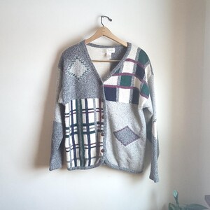 Może przedstawiać: Sweter rozpinany w stylu vintage z patchworkiem. Sweter ma szare, białe, zielone i bordowe kwadraty i romby. Kardigan wisi na drewnianym wieszaku na białej ścianie.