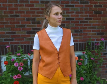 May include: A woman wearing a brown leather vest over a white turtleneck shirt and mustard yellow flared pants. She is standing in front of a brick wall with a window and green grass.