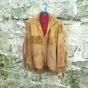 May include: A tan suede jacket featuring long fringe detailing across the chest, shoulders, and sleeves. The jacket has a pointed collar, a central zip closure, and a vibrant red interior lining, displayed on a blue plastic hanger.