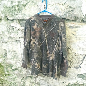May include: A long-sleeve shirt featuring a brown, green, and black camouflage pattern with a forest leaf design. The shirt has a crew neckline, a partial button placket at the front, and solid brown ribbed cuffs at the ends of the long sleeves.