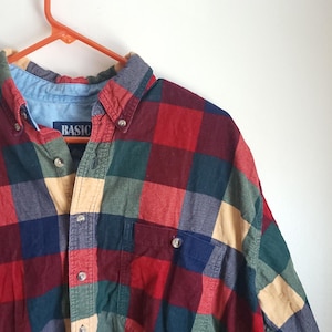 May include: A button-down plaid shirt in red, green, blue, and tan. The shirt has a button-down collar and a chest pocket. The word "BASIC" is visible on the collar.