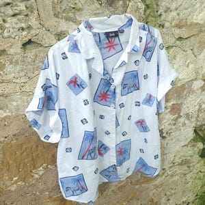 May include: White button-down shirt with a tropical pattern featuring blue squares with palm trees and red accents. The short-sleeved shirt is hanging on a metal hanger against a stone wall.