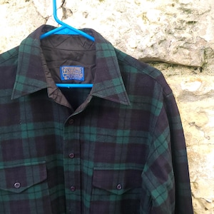 May include: A long-sleeve button-up shirt featuring a dark green and black plaid pattern. The shirt has a pointed collar, two front chest pockets with button flaps, and a blue Pendleton brand label sewn into the inner neck lining.