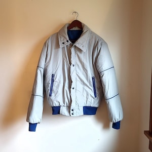 May include: A light gray bomber jacket with blue accents, hanging on a wooden hanger. The jacket features a ribbed blue collar, cuffs, and hem, with blue piping along the seams and two blue zippered pockets. The jacket is buttoned up.
