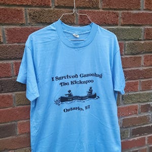 May include: Light blue t-shirt with the text "I Survived Canoeing The Kickapoo" above a graphic of a canoe with two people. Below the graphic is "The Peeling Ceek" and "Ontario, WI". The shirt is hanging on a wire hanger.