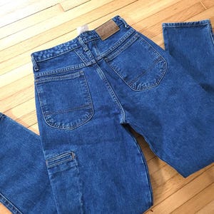 May include: Blue denim jeans with a classic five-pocket design. The jeans have a straight leg cut and a medium wash. The back features two pockets and a leather patch. The jeans are folded on a wooden surface.