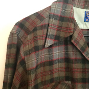 Puede incluir: Primer plano de una camisa a cuadros en tonos marrón, gris y rojo. La camisa tiene un cuello clásico, un bolsillo en el pecho y una etiqueta con texto. La tela tiene un aspecto texturizado.