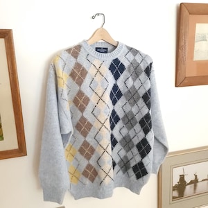 Puede incluir: Un suéter azul claro con diseño de rombos, con cuello redondo, colgado de una percha de madera. El patrón de rombos presenta diamantes en marrón, amarillo, azul marino y gris. El suéter se exhibe contra una pared blanca con obras de arte enmarcadas.