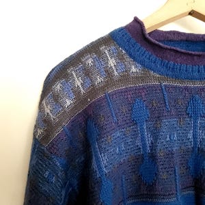 May include: A close-up of a vintage sweater in shades of blue, purple, and gray. The sweater features a geometric pattern on the shoulders and a ribbed collar. The garment is hanging on a wooden hanger against a white background.