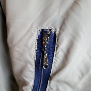 May include: Close-up of a jacket detail featuring a silver zipper with a blue interior. The jacket is a light beige color, with the zipper pull showing a decorative design. The zipper is partially open, revealing the blue fabric beneath.