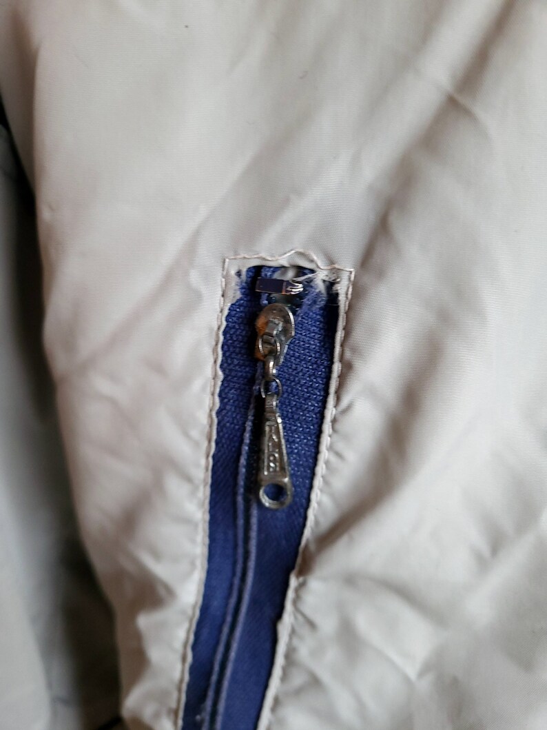 May include: Close-up of a jacket detail featuring a silver zipper with a blue interior. The jacket is a light beige color, with the zipper pull showing a decorative design. The zipper is partially open, revealing the blue fabric beneath.