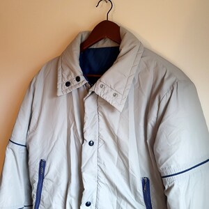May include: A light gray jacket with a blue interior and trim, hanging on a wooden hanger. The jacket has a button-up front, two zippered pockets, and a high collar. The jacket appears to be made of a lightweight, possibly water-resistant material.