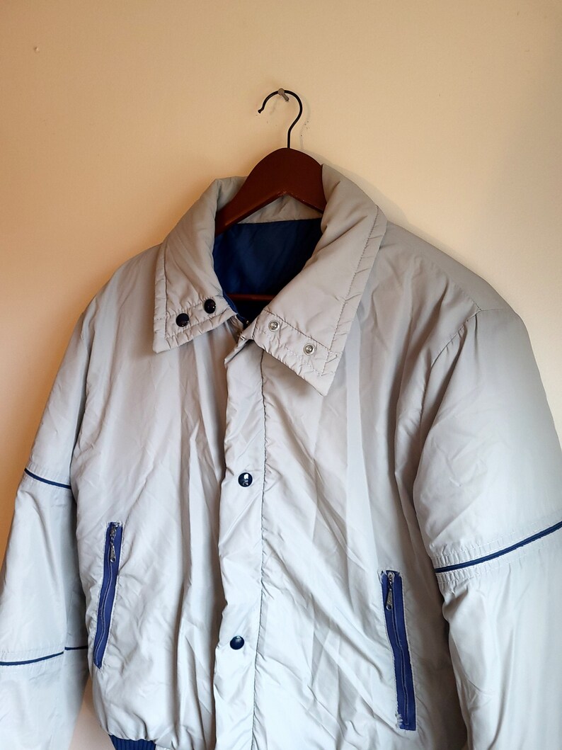 May include: A light gray jacket with a blue interior and trim, hanging on a wooden hanger. The jacket has a button-up front, two zippered pockets, and a high collar. The jacket appears to be made of a lightweight, possibly water-resistant material.
