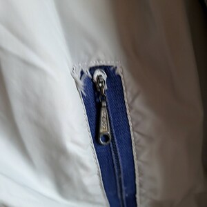 May include: Close-up of a light gray jacket with a blue zipper and trim. The zipper pull has the word "ZION" printed on it. The fabric is slightly torn near the zipper.