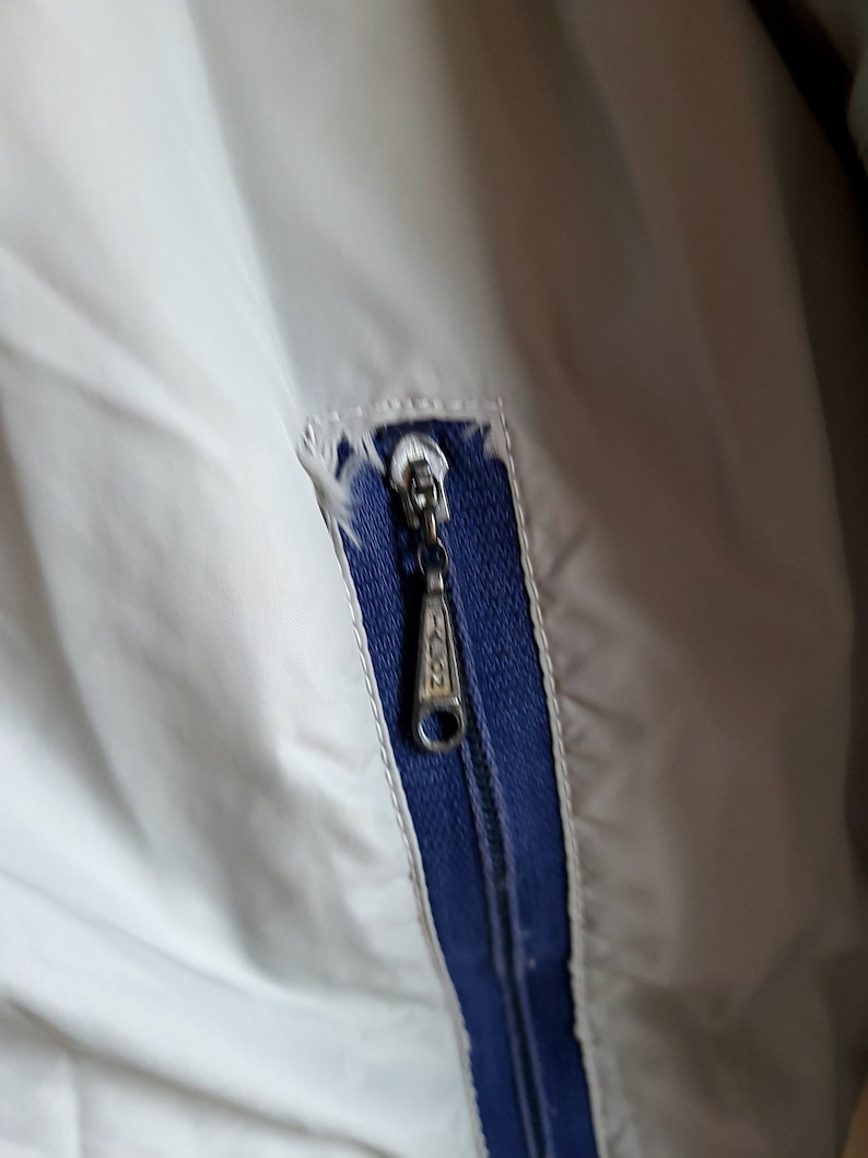 May include: Close-up of a light gray jacket with a blue zipper and trim. The zipper pull has the word "ZION" printed on it. The fabric is slightly torn near the zipper.