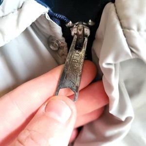 May include: Close-up of a silver metal zipper on a jacket. The zipper pull is rectangular with a textured surface and the word "IDEAL" is visible. The jacket has a white and blue color scheme.