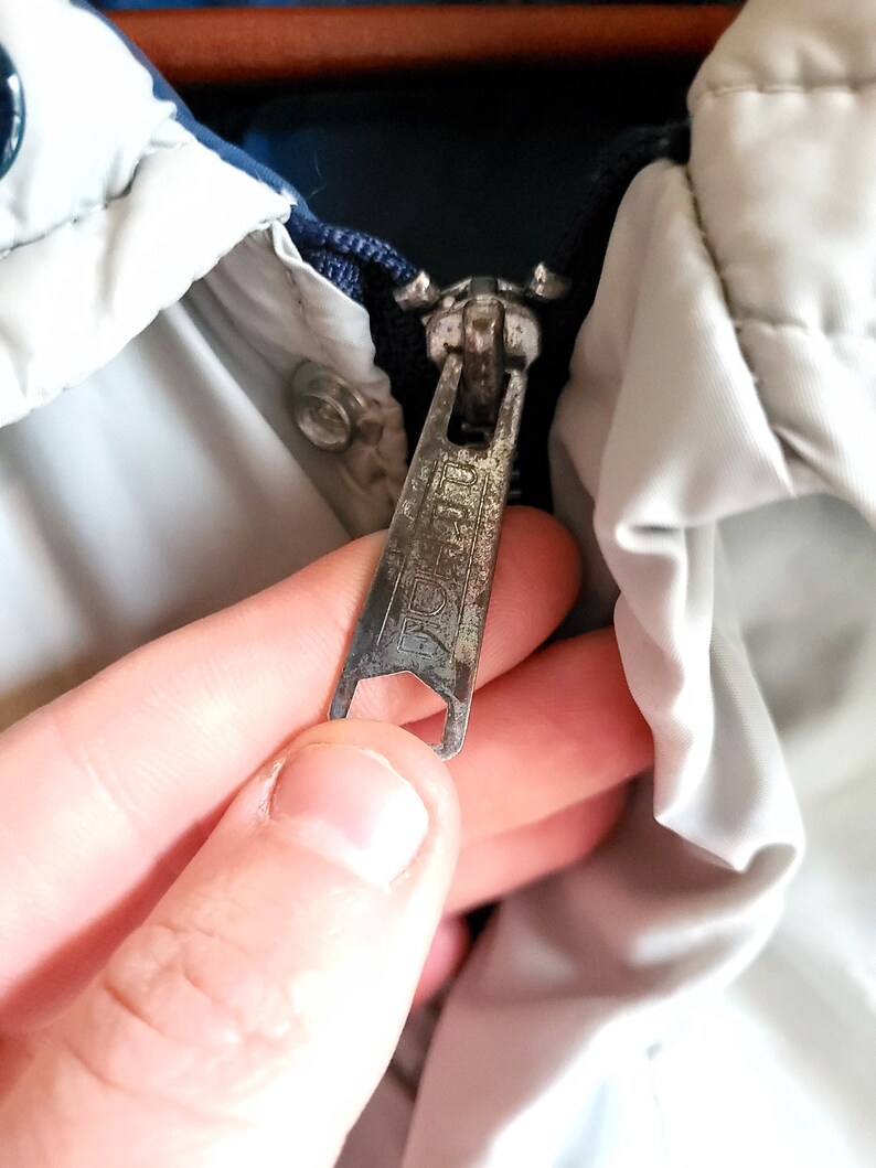 May include: Close-up of a silver metal zipper on a jacket. The zipper pull is rectangular with a textured surface and the word "IDEAL" is visible. The jacket has a white and blue color scheme.