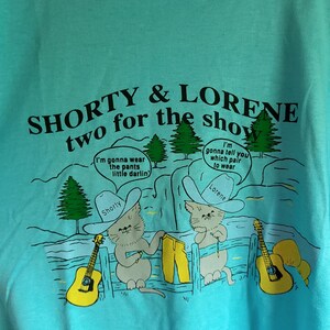 May include: A turquoise blue t-shirt with a cartoon image of two cats wearing cowboy hats and holding guitars. The text on the shirt reads "SHORTY & LORENE two for the show".