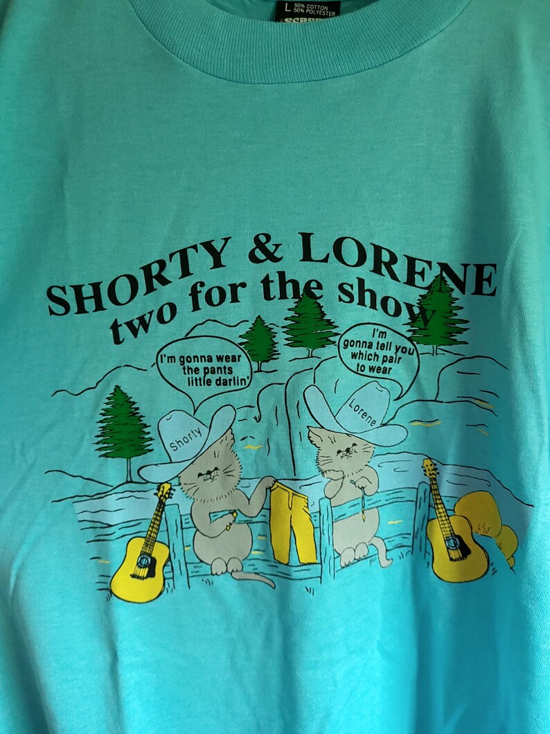 May include: A turquoise blue t-shirt with a cartoon image of two cats wearing cowboy hats and holding guitars. The text on the shirt reads "SHORTY & LORENE two for the show".