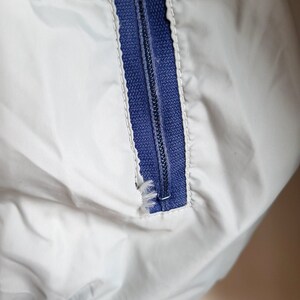 May include: Close-up of a white jacket with a blue zipper. The zipper is partially closed, revealing the teeth and the blue fabric underneath. The jacket has a blue ribbed cuff.