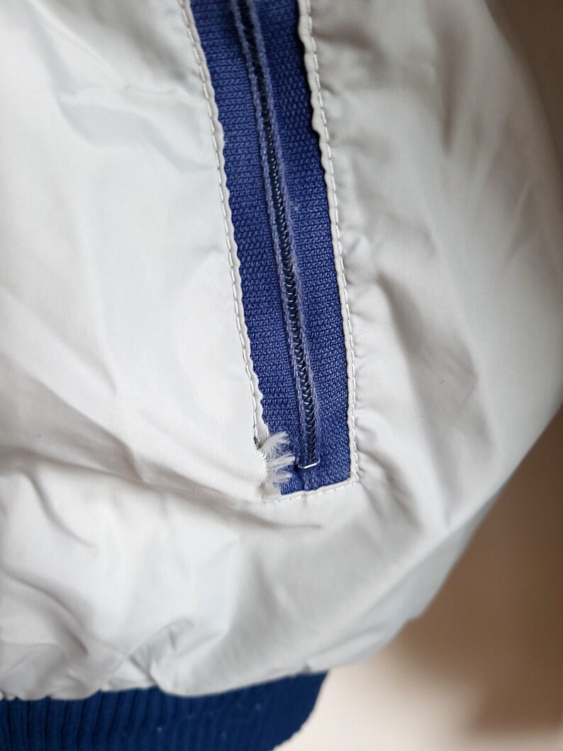 May include: Close-up of a white jacket with a blue zipper. The zipper is partially closed, revealing the teeth and the blue fabric underneath. The jacket has a blue ribbed cuff.