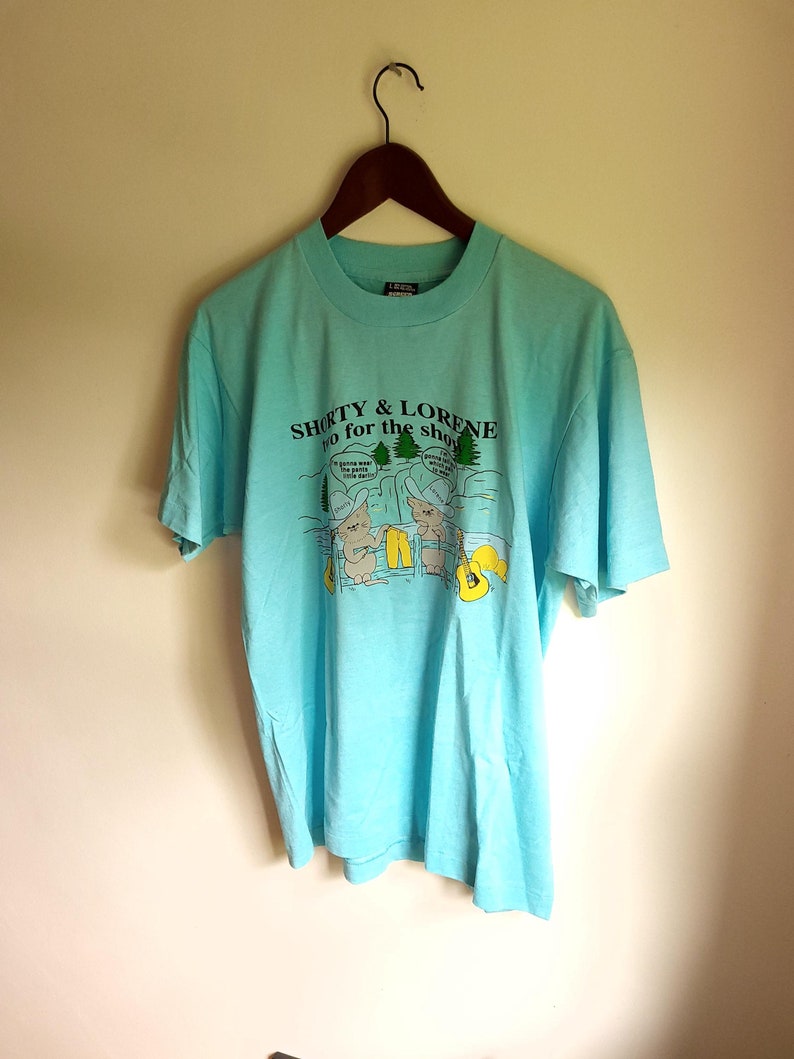 May include: A light blue t-shirt with a cartoon graphic of two people fishing. The text on the shirt reads "Shorty & Lorene Go for the  Shroom".