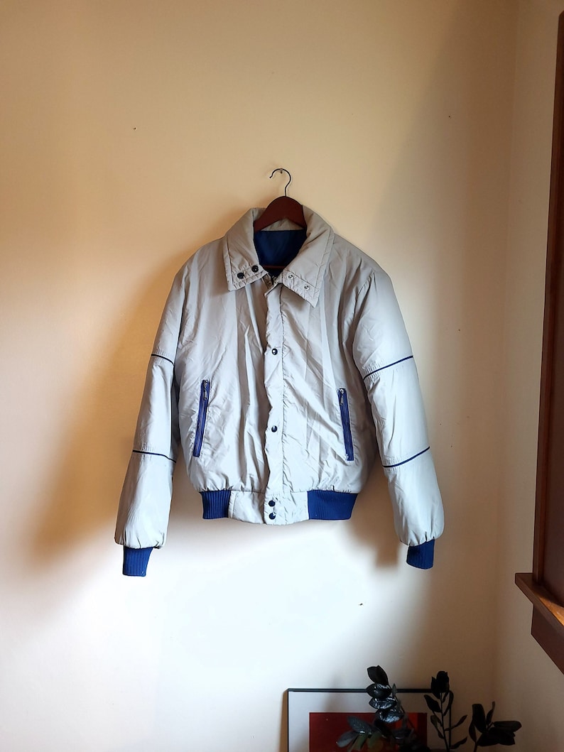 May include: A light gray bomber jacket with blue accents, hanging on a wooden hanger. The jacket features a ribbed blue collar, cuffs, and hem, with blue piping along the seams and two blue zippered pockets. The jacket is buttoned up.