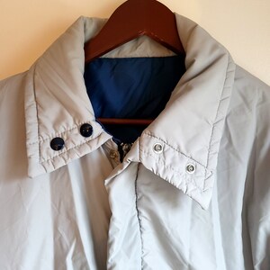 May include: A light gray reversible jacket with a blue interior, hanging on a wooden hanger. The jacket has a high collar, snap closures, and a zipper. The jacket is a neutral color and appears to be lightly padded.