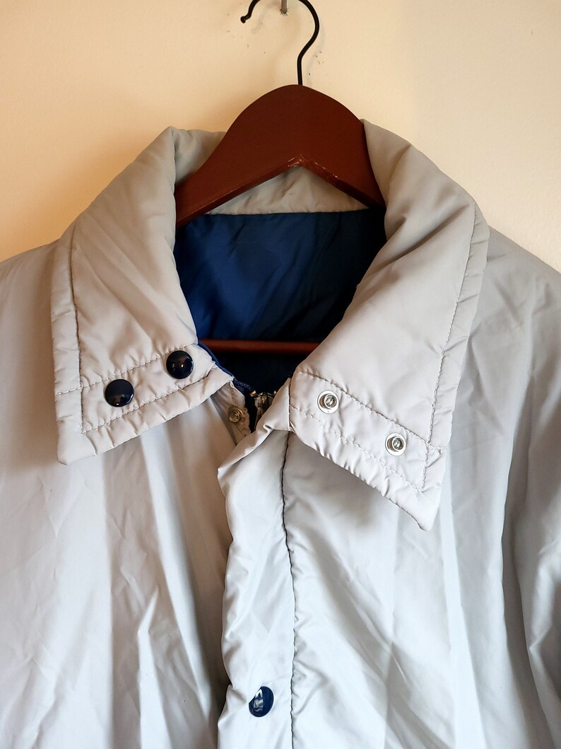 May include: A light gray reversible jacket with a blue interior, hanging on a wooden hanger. The jacket has a high collar, snap closures, and a zipper. The jacket is a neutral color and appears to be lightly padded.