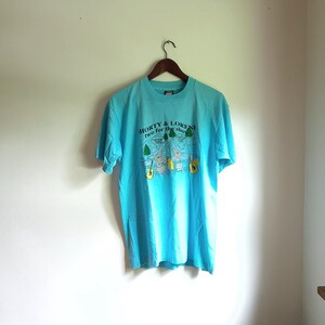 May include: A turquoise blue t-shirt with a graphic print of two cartoon figures holding guitars. The text on the shirt reads "Shorty & Lorene two for the show".