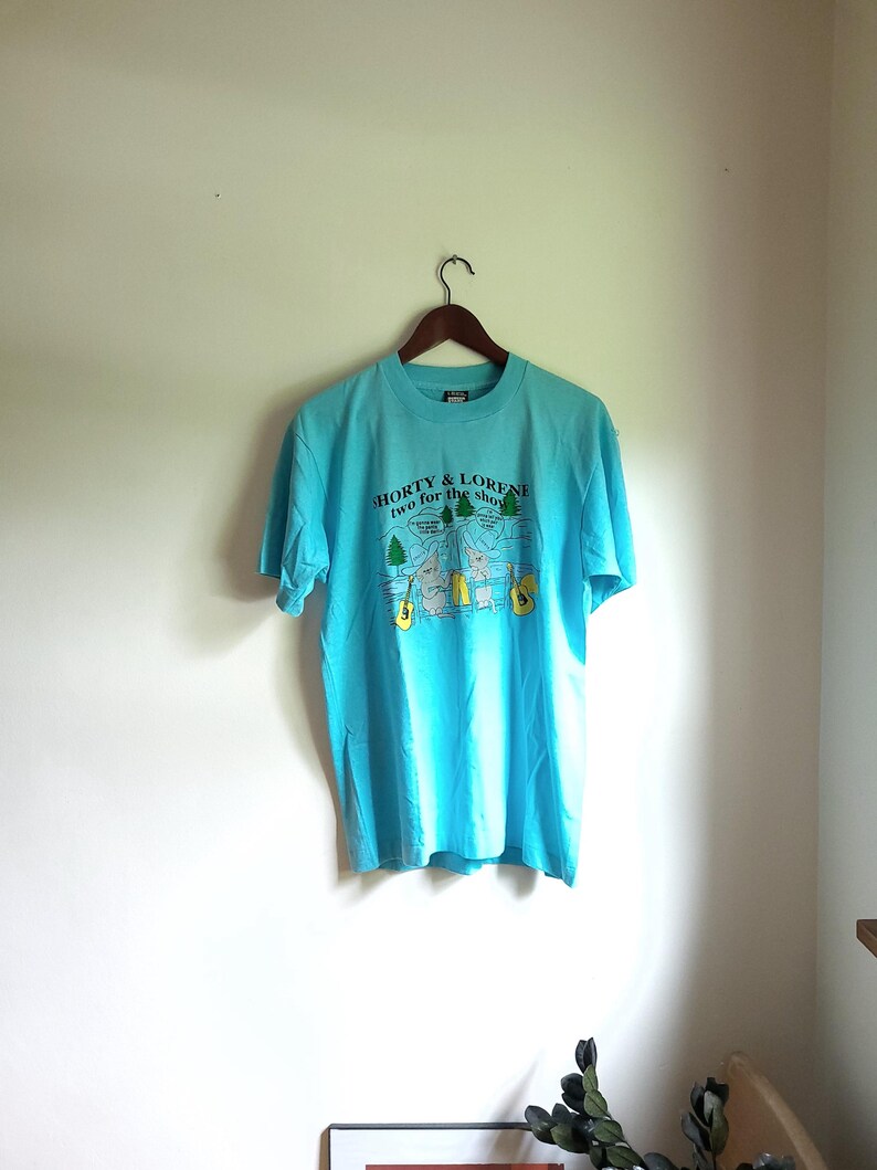May include: A turquoise blue t-shirt with a graphic print of two cartoon figures holding guitars. The text on the shirt reads "Shorty & Lorene two for the show".