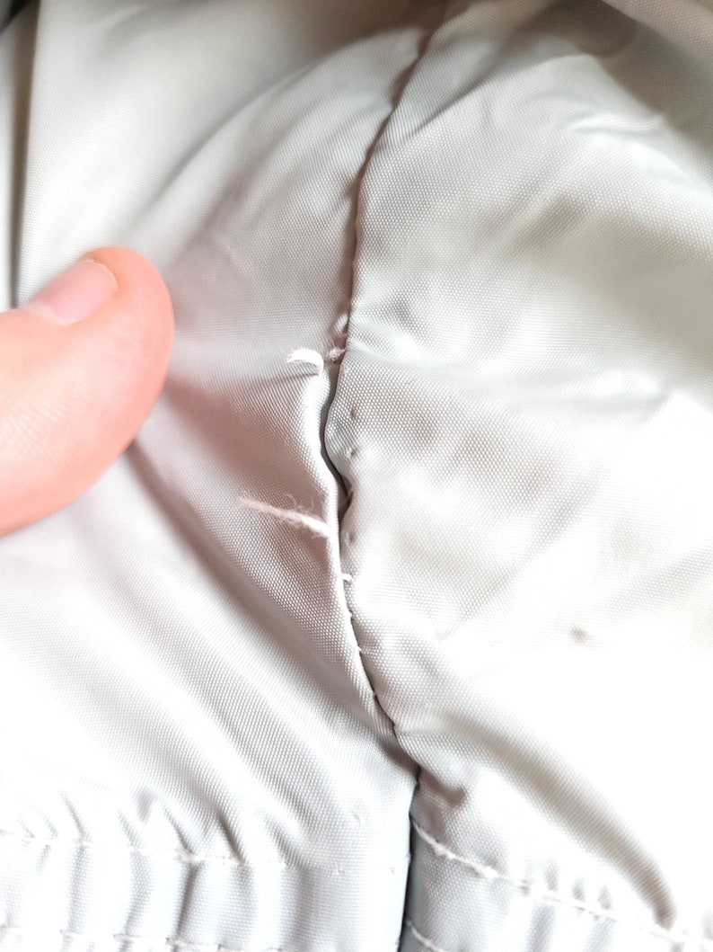 May include: Close-up of a light gray fabric with a visible seam and small tears. The fabric appears to be a smooth, possibly synthetic material. The seam shows signs of wear and tear, with loose threads.