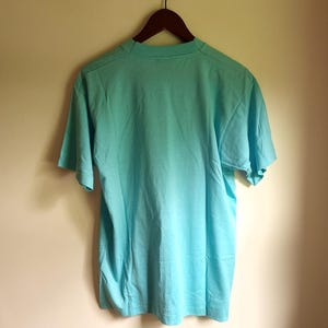 May include: A light blue t-shirt hanging on a wooden hanger against a white wall.