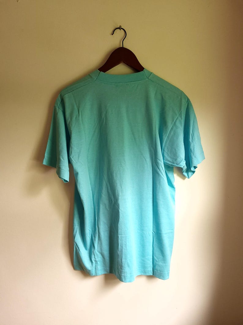 May include: A light blue t-shirt hanging on a wooden hanger against a white wall.