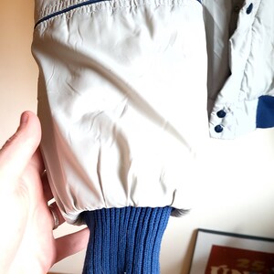 May include: Close-up of a light gray jacket sleeve with a navy blue ribbed knit cuff. The jacket has a navy blue trim along the seams and a button closure. The sleeve is held by a hand.