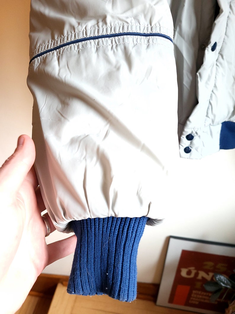 May include: Close-up of a light gray jacket sleeve with a navy blue ribbed knit cuff. The jacket has a navy blue trim along the seams and a button closure. The sleeve is held by a hand.